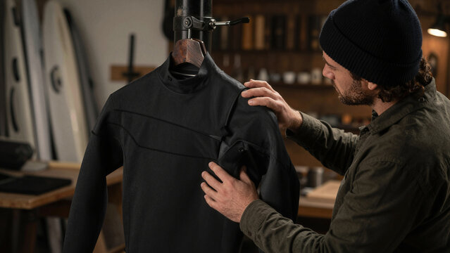 A person looks closely at a black wetsuit in a workshop. They are checking the features of the suit on a wooden hanger. This scene takes place indoors, with surfboards in the background