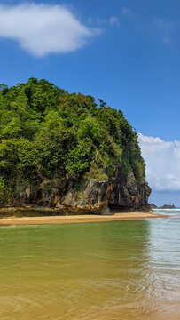 Vertical view of a green covered rocky cliff beside calm estuarine waters meeting ocean waves at Taman Beach in Pacitan, East Java, Indonesia.