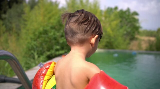 Happy toddler wearing inflatable arm floaties turns and smiles by a pool edge, pretending to jump into the water