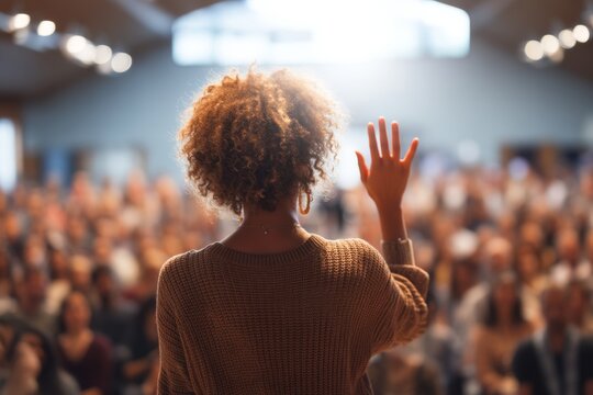 Confident speaker addressing a large audience with raised hand in a well-lit venue showcasing public speaking and community engagement. Generative AI