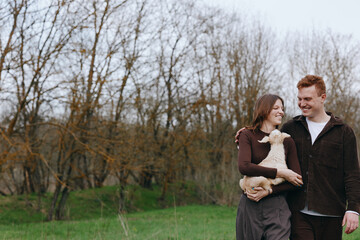 Naklejka premium Happy couple outdoors with a dog, smiling and affectionate, walking through a park in bright daylight, enjoying nature and each other, expressing warmth and companionship on a calm day.