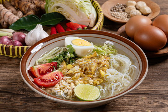 Authentic indonesian chicken soto soup in brown rimmed bowl featuring glass noodles lime egg and raw spices