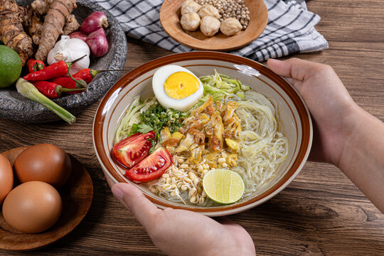 Hands holding authentic indonesian chicken soto soup bowl featuring glass noodles boiled egg and traditional spices background