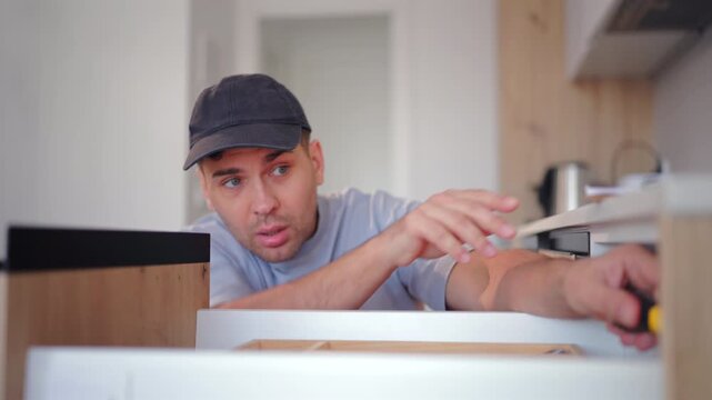 Handyman with toolbox performing maintenance in kitchen of home for repair. Young man wearing cap uses screwdriver to fix cabinet drawer in modern domestic interior with wooden accents.
