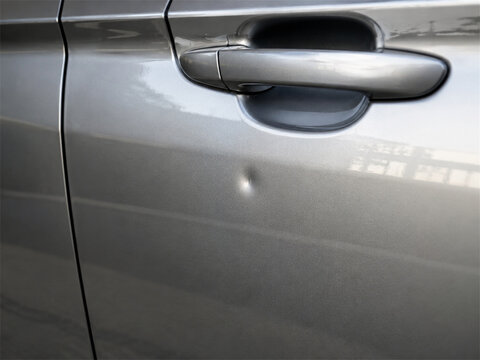 Close view of a silver car door with a small parking dent below the handle showing minor body damage on metallic paint before professional paintless dent repair service and restoration work