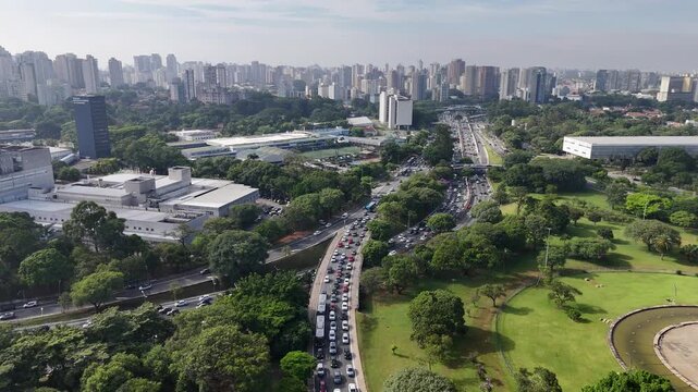 Aerial view of bustling highway traffic next to Ibirapuera Park with the Obelisk and Sao Paulo skyline in the background.