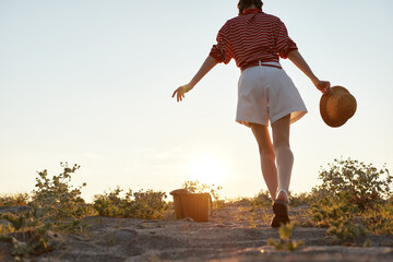 Fototapeta premium A young woman in a striped shirt and white shorts walks toward a golden sunset on a sandy path, her hat swinging in a relaxed, carefree summer moment.