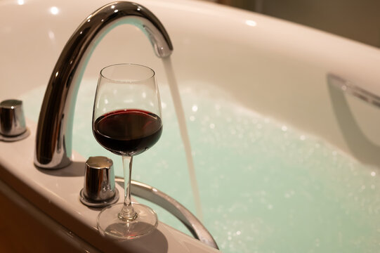 Glass of red wine by a filling bathtub with bubble foam.