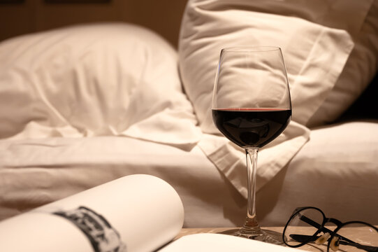 Glass of red wine, book, and glasses on a table in front of a bedroom bed.