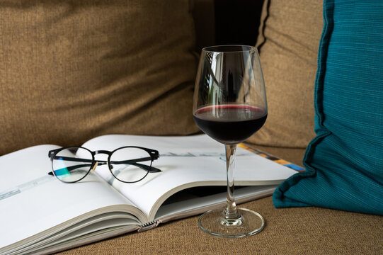 Glass of red wine, book, and glasses on a sofa with cushions.