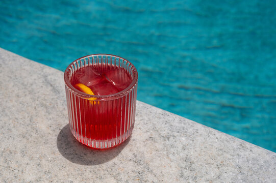 Red cocktail with ice and lemon slice by the swimming pool edge.
