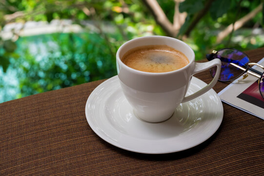 Cup of hot coffee with a saucer, book, and glasses on a table in a garden.