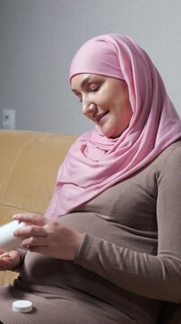 Young pregnant muslim woman in a pink hijab sits on the sofa, carefully reading the information on a bottle of prenatal vitamins, prioritizing her baby's health and her well being