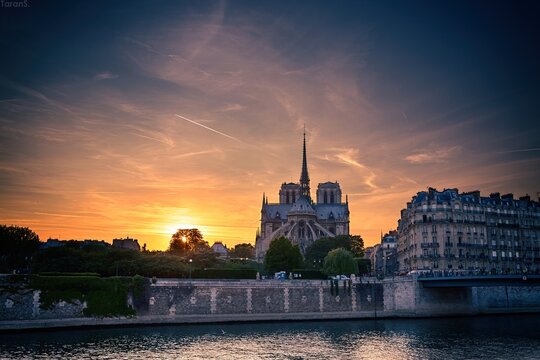 Notre Dame cathedral silhouette against dramatic orange sunset sky with Seine river and Parisian buildings in foreground, France landmark scene
