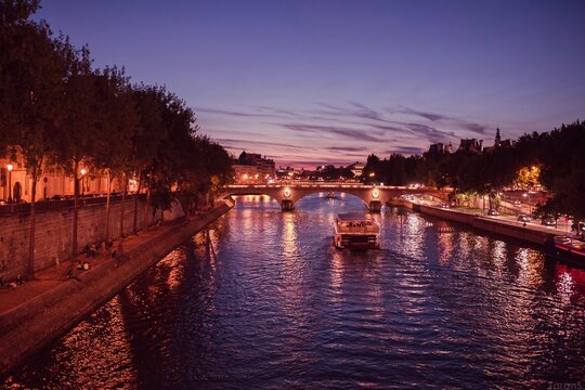 Seine river bridge illuminated at sunset with tour boat passing beneath, Paris embankment and city skyline reflected in water, France evening scene