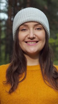 Close up portrait of an attractive woman in a beanie hat and yellow sweater. Her expression slowly transitions from serious to a bright, happy smile, looking directly at the camera in a park
