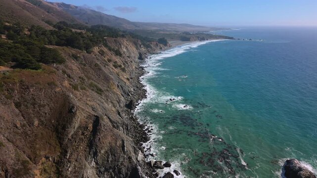 Elevated view of rugged coastal cliffs descending to rocky shoreline where turquoise shallow water, submerged stones, kelp beds, and foamy surf flow into deeper blue ocean beneath clear bright sky