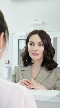 Young businesswoman with dark hair, dressed in a stylish jacket, engaging in a conversation with a female receptionist at a modern clinic's reception desk, actively discussing services