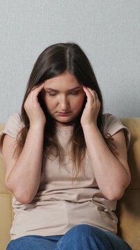 Depressed young woman sitting on a couch, feeling intense stress and massaging her temples due to a severe headache, suffering from migraine, anxiety, and mental health issues