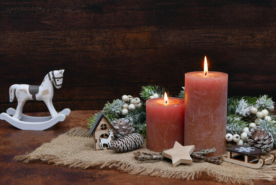 A Christmas still life featuring two lit dusky pink candles, a rocking horse, and decorations on jute against a wooden wall with space for text.
