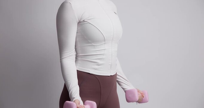 Slow motion woman performing dumbbell bicep curls in indoor studio with plain backdrop and neutral lighting.