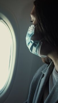 Young female passenger wearing a protective face mask sitting inside an aircraft and looking out the window, a concept of safe air travel and tourism during a global health crisis