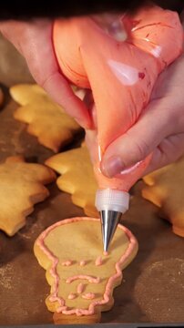 Baker's hands expertly decorating freshly baked skull shaped sugar cookies with delicate pink royal icing, carefully adding details to the spooky and sweet halloween treats
