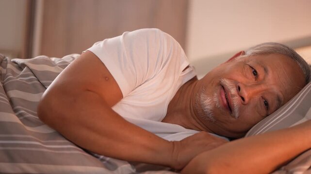 Elderly man lies awake on his pillow and breaks into a slow contented smile in the early morning light. 4K senior healthcare footage shot in a modern Asian hospital room.