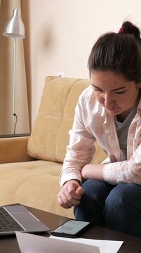 Worried woman sitting on a couch calculating expenses on her smartphone and getting frustrated by financial problems, covering her face in despair after seeing bills and debts