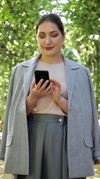 Cheerful plus size businesswoman with professional makeup using her mobile phone while standing in a beautiful park, enjoying a moment of relaxation and staying connected online