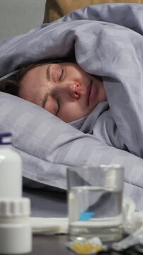 Young ill woman sleeping in bed at home, covered in a blanket and suffering from a cold, flu, or coronavirus, coughing heavily with medicines and a glass of water on the nightstand