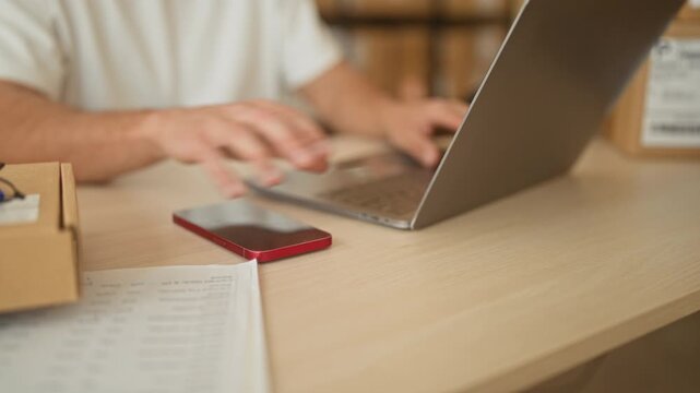 Man holding smartphone and typing on laptop in a building, surrounded by parcels and a notebook; focused entrepreneurship.