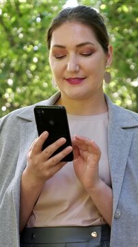 Professional businesswoman in a gray jacket using her smartphone in a city park, elegantly scrolling through her social media feed and smiling while checking messages on a sunny day