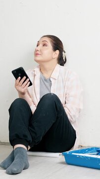 Relaxed young woman sitting on the floor next to a paint tray, taking a well deserved break from diy home repairs and browsing social media or online stores on her smartphone