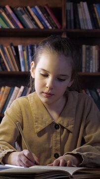 Concentrated young girl writing with a pencil in a notebook while sitting at a desk. A focused student doing her homework in a library, surrounded by bookshelves under a warm light