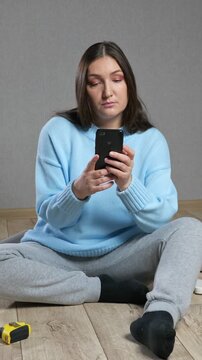 Thoughtful woman sitting on the floor in an empty room, using a smartphone application for home renovation, measuring walls, and choosing furniture for her new apartment