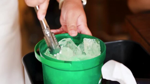 Hand Using Metal Tongs to Pick Ice from Green Bucket