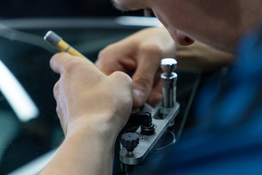 Professional auto mechanic carefully repairing chipped car windshield glass in workshop, concentrated technician using specialized tools for vehicle windscreen restoration inside service garage.
