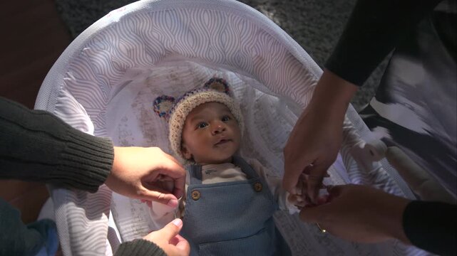 African American young family gently holding tiny hands of their newborn baby lying in white bassinet wearing knitted bear hat and blue overalls at home in soft warm daylight.