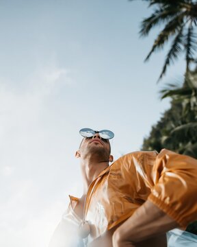 Man in a glossy orange rain jacket and mirrored sunglasses looking upward against a tropical sky, for streetwear and travel advertising with copy space
