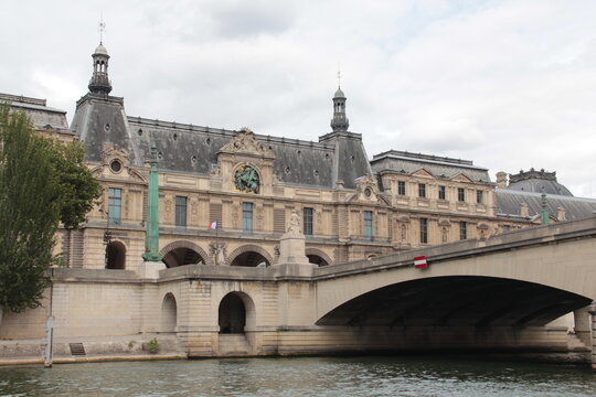 A moody view of the Seine River bordered by stone embankments and historic Parisian buildings, with a bridge connecting both sides under gray clouds.