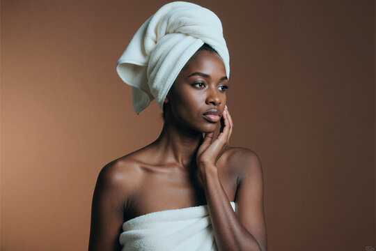 Young adult Black woman touching cheek after skincare routine, wrapped in towel and presenting self care beauty concept. Ideal for spa, skincare, wellness, cosmetic branding, medium shot