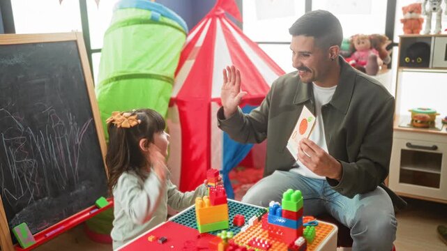 Man raises hand for high five while holding a book and guiding girl with blocks at a table in a playroom inside building; playful learning.