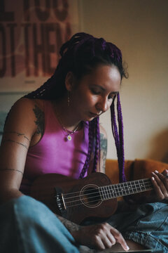Tattooed woman with purple braids learning ukulele at home