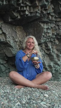 Blonde woman playing singing bowl while sitting cross-legged on pebble beach. Vertical medium shot.