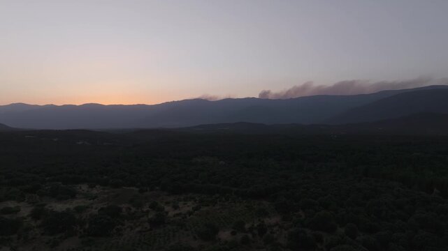 Ascending drone view at sunset over the Central Mountain Range, revealing a large smoke plume and a smaller column emerging beyond the mountain ridges in the Burgohondo area.
