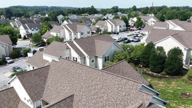Dense townhome cluster with matching tan gabled roofs and white siding fills modern American neighborhood, driveways and parking lots wind between houses and mature trees. Aerial drone rising wide