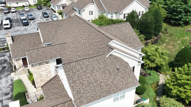 Steep asphalt shingle roof of upscale townhome dominates frame beside parking lot, mature shrubs and mulched garden beds fill quiet suburban American neighborhood, USA. Aerial drone orbit shot.