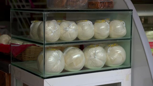 Static medium shot of traditional Mexican Queso Oaxaca balls packed in plastic bags inside a glass display case at a local store.