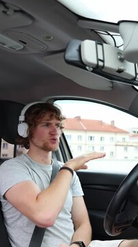 Vertical screen driver listens to music through headphones and dances with energetic gestures inside a parked car enjoying carefree entertainment while staying stationary